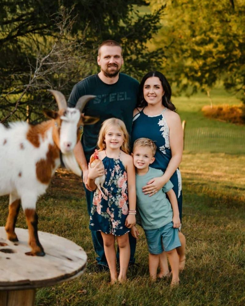 A family of four stands outdoors with two children, flanked by a curious goat. They are smiling, surrounded by greenery in a rustic setting.